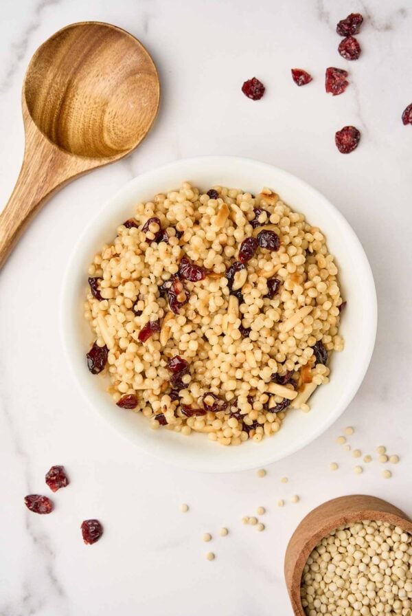 A bowl of toasted pearl couscous with dried cranberries and slivered almonds surrounded by a wooden spoon, scattered cranberries, and a wooden measuring cup with uncooked Israeli couscous.