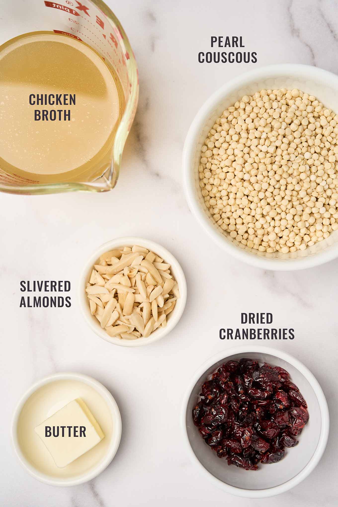A labeled, overhead photo of the ingredients needed to make pearl couscous with cranberries and almonds; pearl couscous, chicken broth, butter, slivered almonds, and dried cranberries.