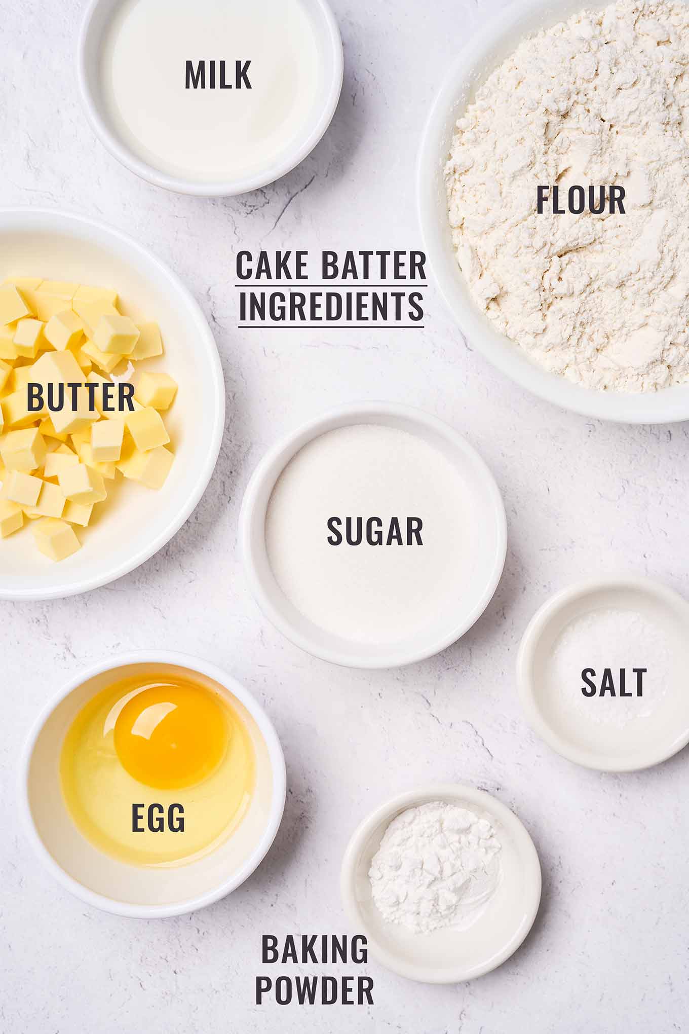 Labeled, overhead view of the ingredients for Golden Delicious apple cake batter in prep bowls; including flour, butter, sugar, egg, milk,baking powder, and salt.
