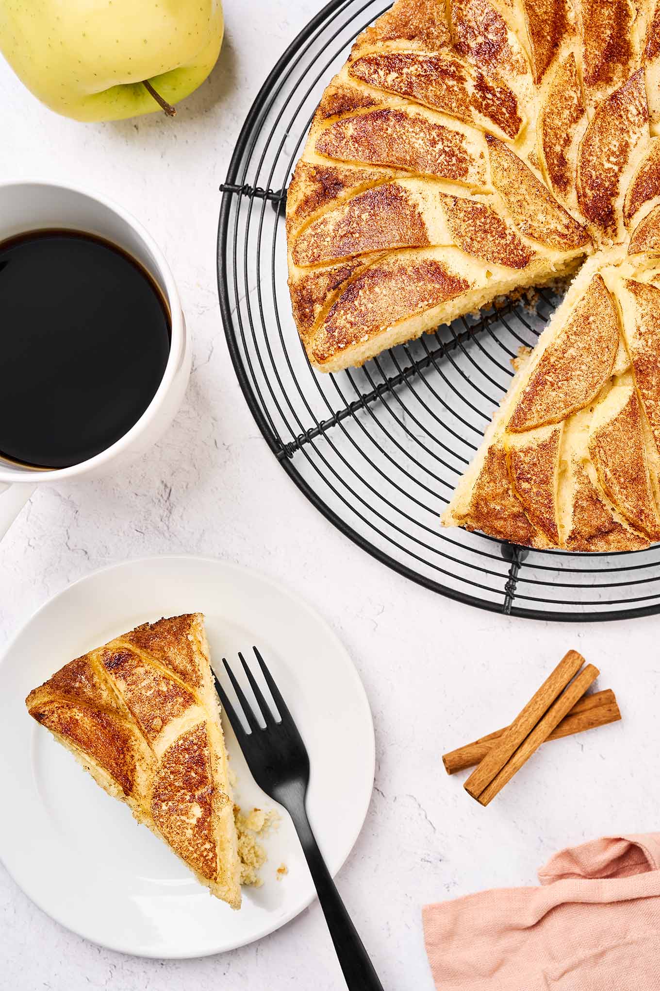 A slice of Golden Delicious apple cake served with a cup of black coffee more cake to the right, cinnamon sticks and a whole apple in the background.