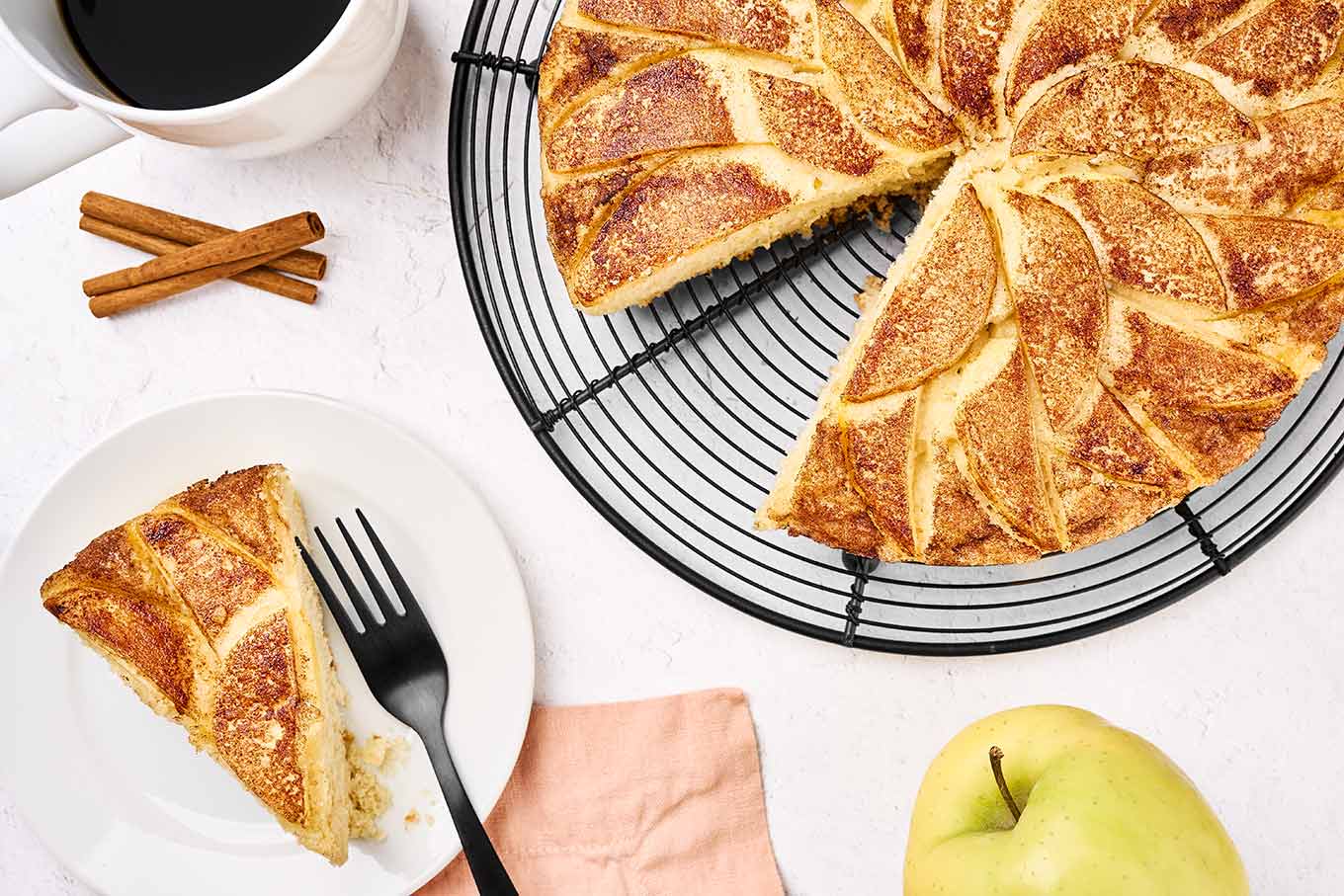 A slice of apple cake on a plate with a black fork, a cup of coffee in the corner, and the rest of the cake on a black wire cooling rack.