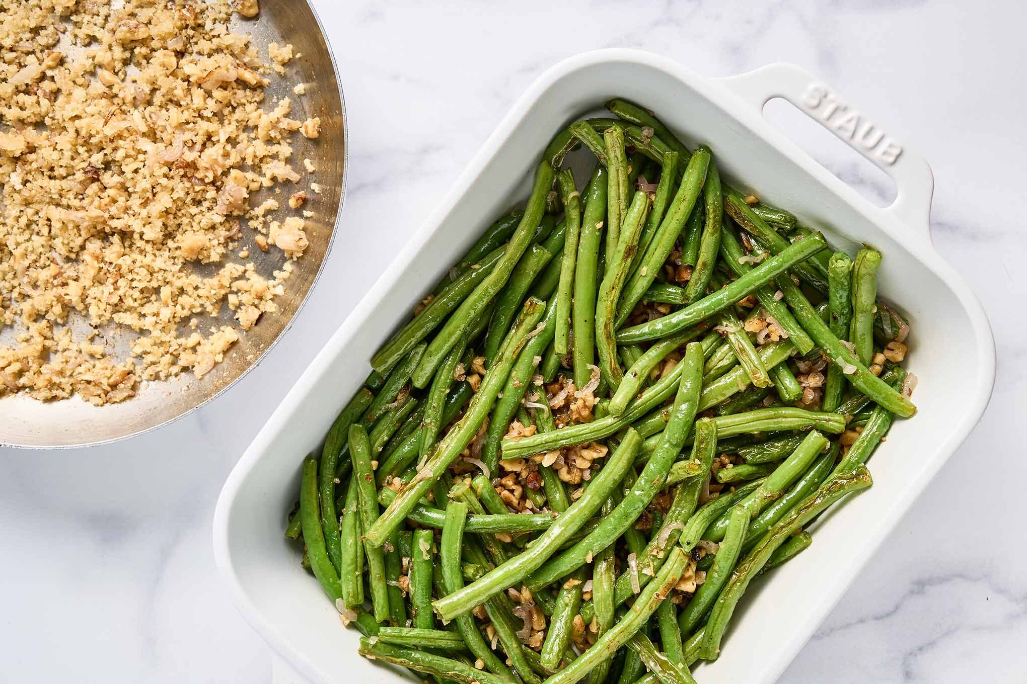 Roasted green beans mixed with saut&eacute;ed shallots and walnuts in a white baking dish, with panko and walnut topping in a skillet beside it.