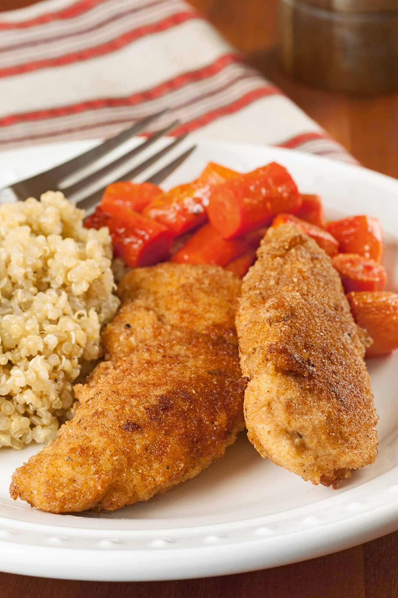 Two spicy breaded chicken tenderloins on a plate with glazed carrots and quinoa, a fork and kitchen towel in the background.