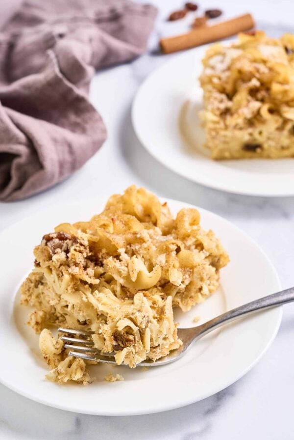 Slice of sweet noodle kugel on a white plate with a fork, showing its crisp cinnamon topping and tender egg noodles.