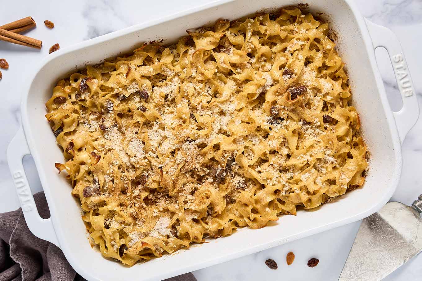 Sweet noodle kugel baked in a white casserole dish with a golden cinnamon topping and visible raisins.