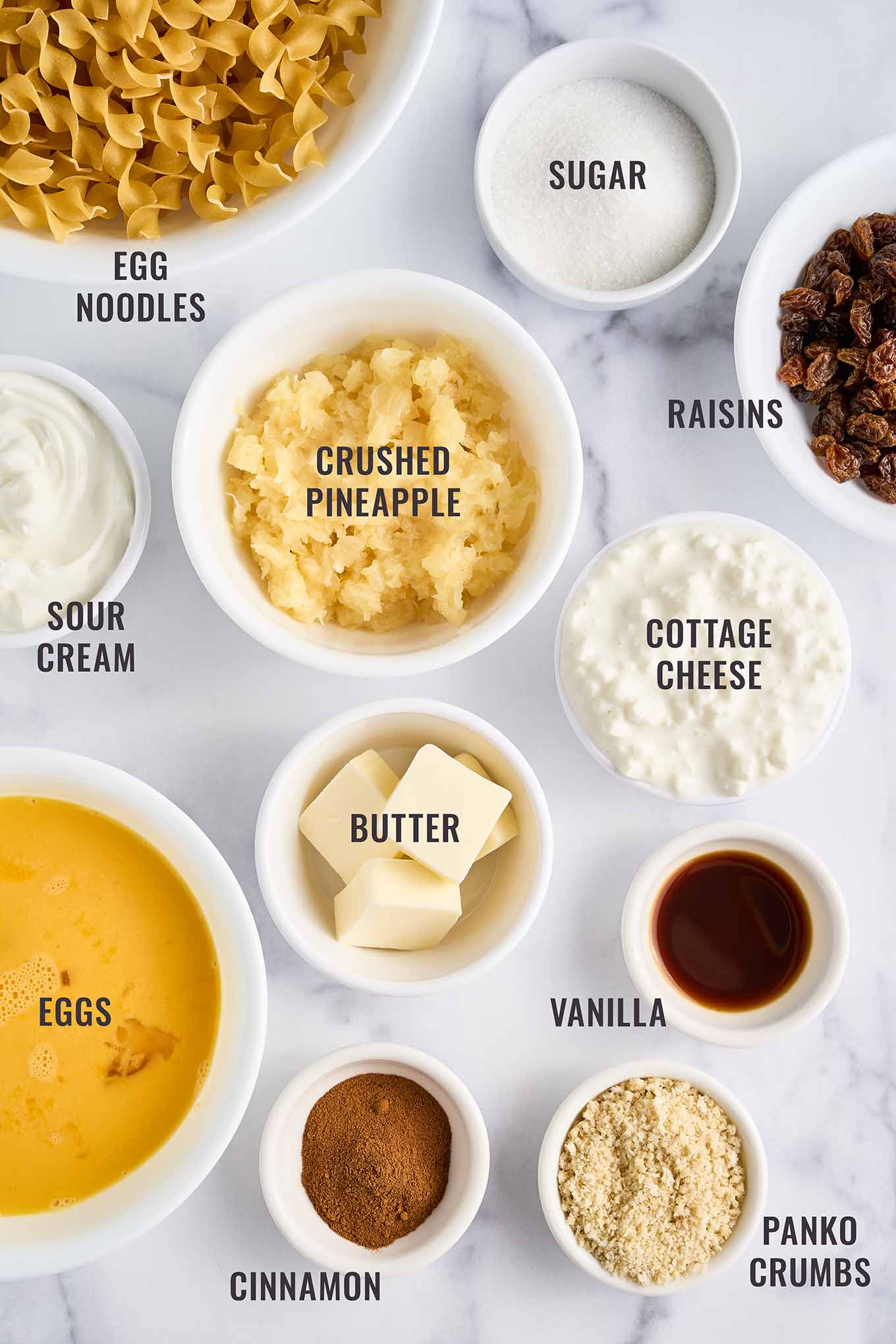 Labeled ingredients for sweet noodle kugel arranged on a marble surface, including egg noodles, crushed pineapple, cottage cheese, sour cream, raisins, eggs, butter, cinnamon, sugar, vanilla, and panko crumbs.