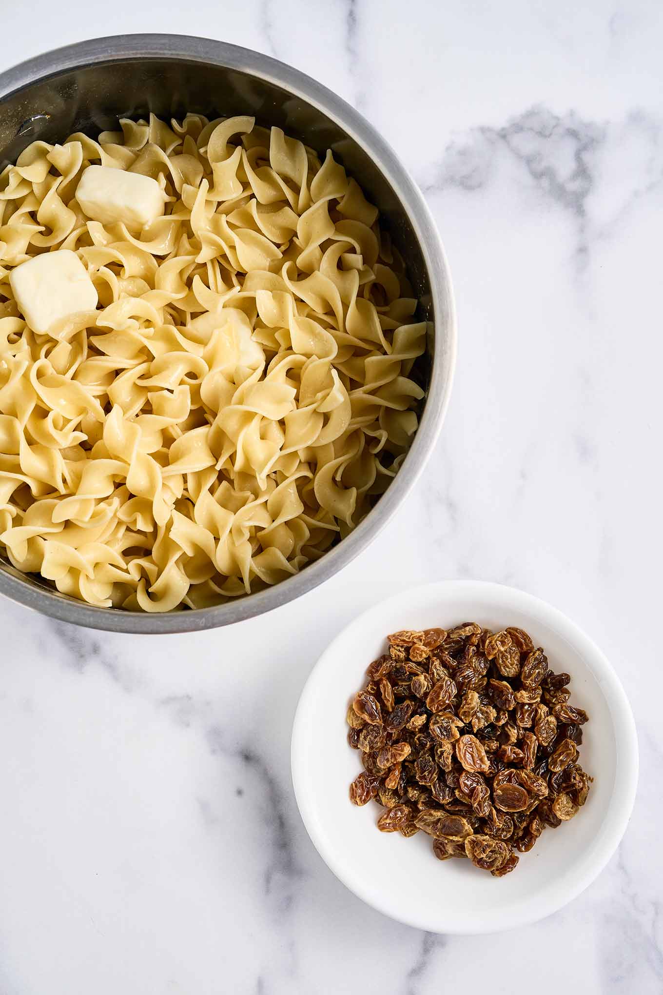Cooked egg noodles with butter in a mixing bowl alongside a small bowl of plumped raisins.