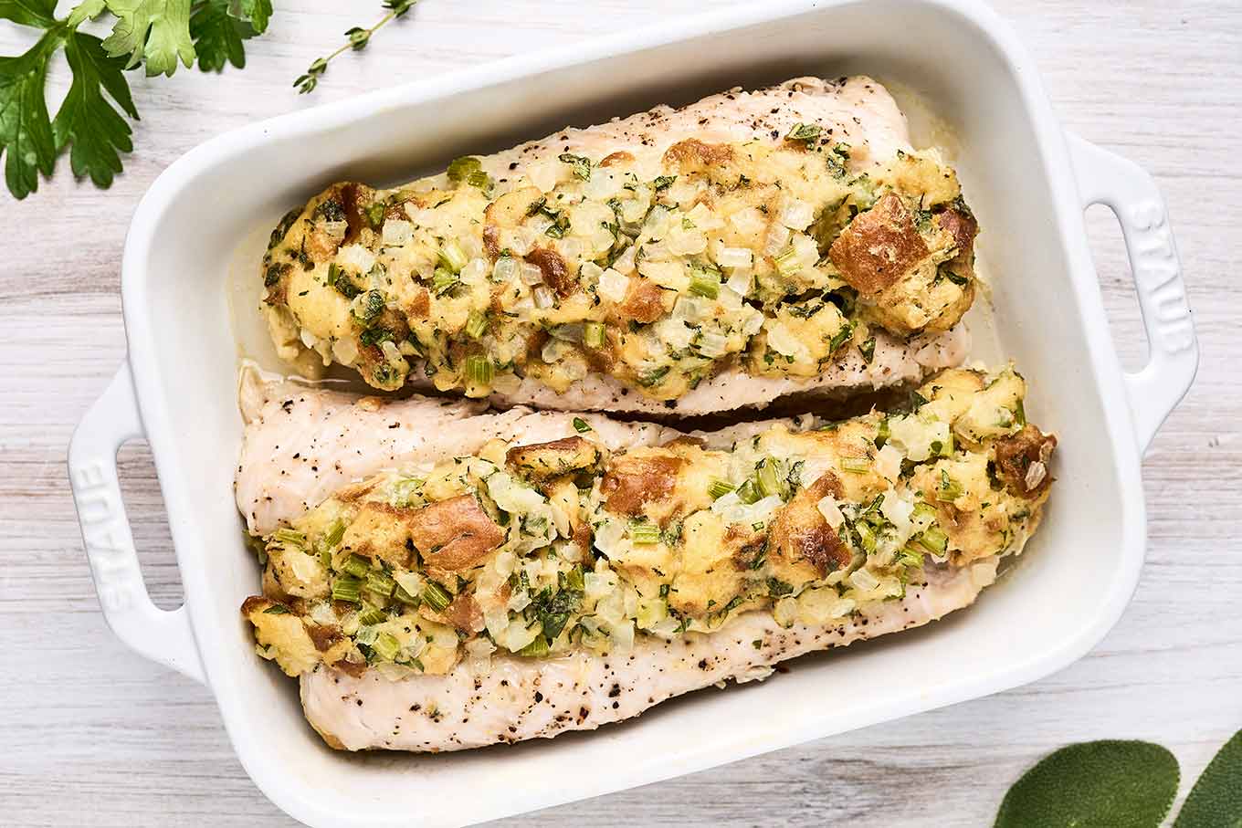 Two stuffed turkey tenderloins in a white oblong baking dish surrounded by fresh parsley, sage and thyme.