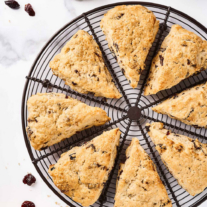 Cherry scones on a round, black cooling rack surrounded by scattered dried cherries, a pitcher of buttermilk, half a navel orange, and a measuring spoon.