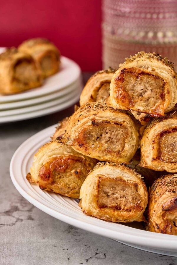 Puff pastry sausage rolls with golden, flaky layers and seasoned sausage filling shown stacked on a white serving plate.