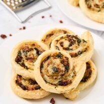 Golden spinach pinwheels made with puff pastry, spinach, sun-dried tomatoes, and cheese, stacked on a white plate.