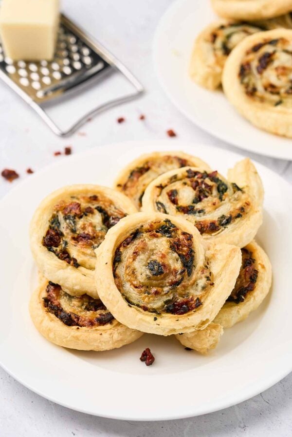 Golden spinach pinwheels made with puff pastry, spinach, sun-dried tomatoes, and cheese, stacked on a white plate.