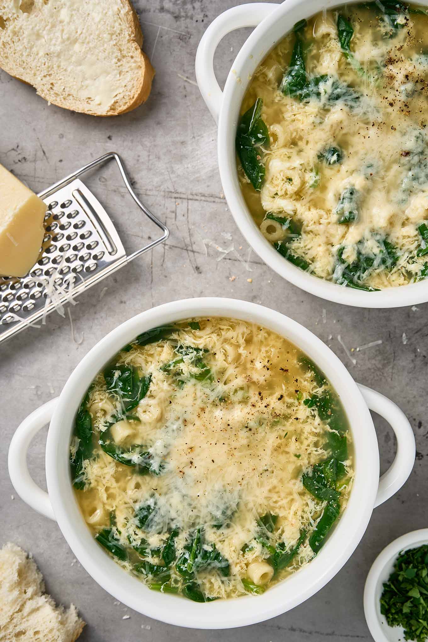 Bowls of stracciatella soup with spinach, ditalini, and grated Parmigiano-Reggiano, shown with bread and a cheese grater.