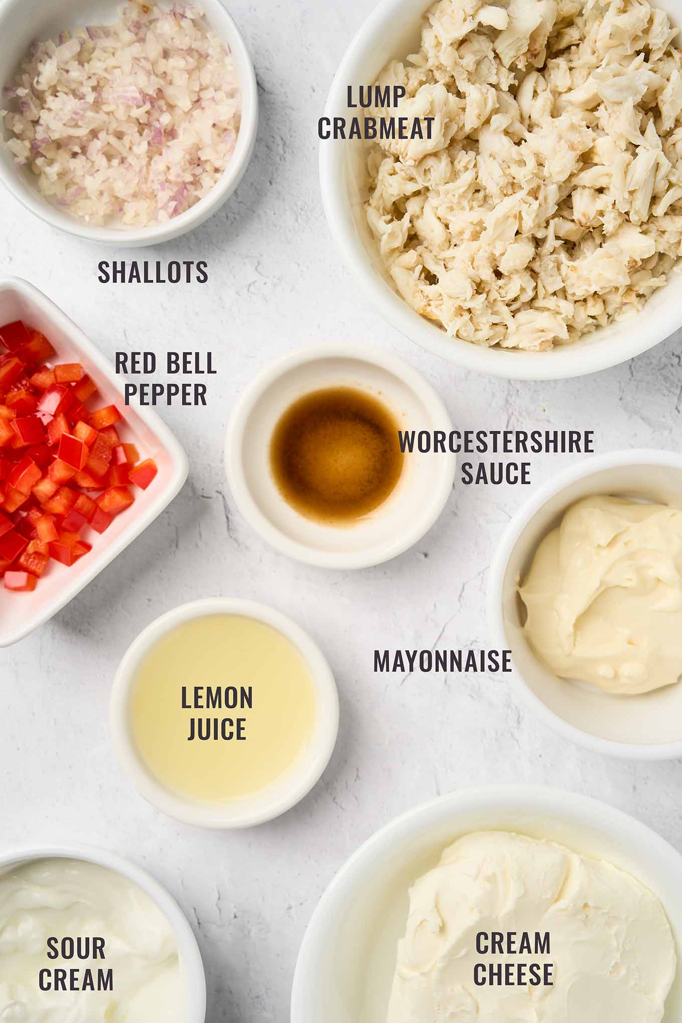 Labeled ingredients for warm crab dip arranged on a light surface, including lump crabmeat, shallots, red bell pepper, lemon juice, Worcestershire sauce, mayonnaise, sour cream, and cream cheese.