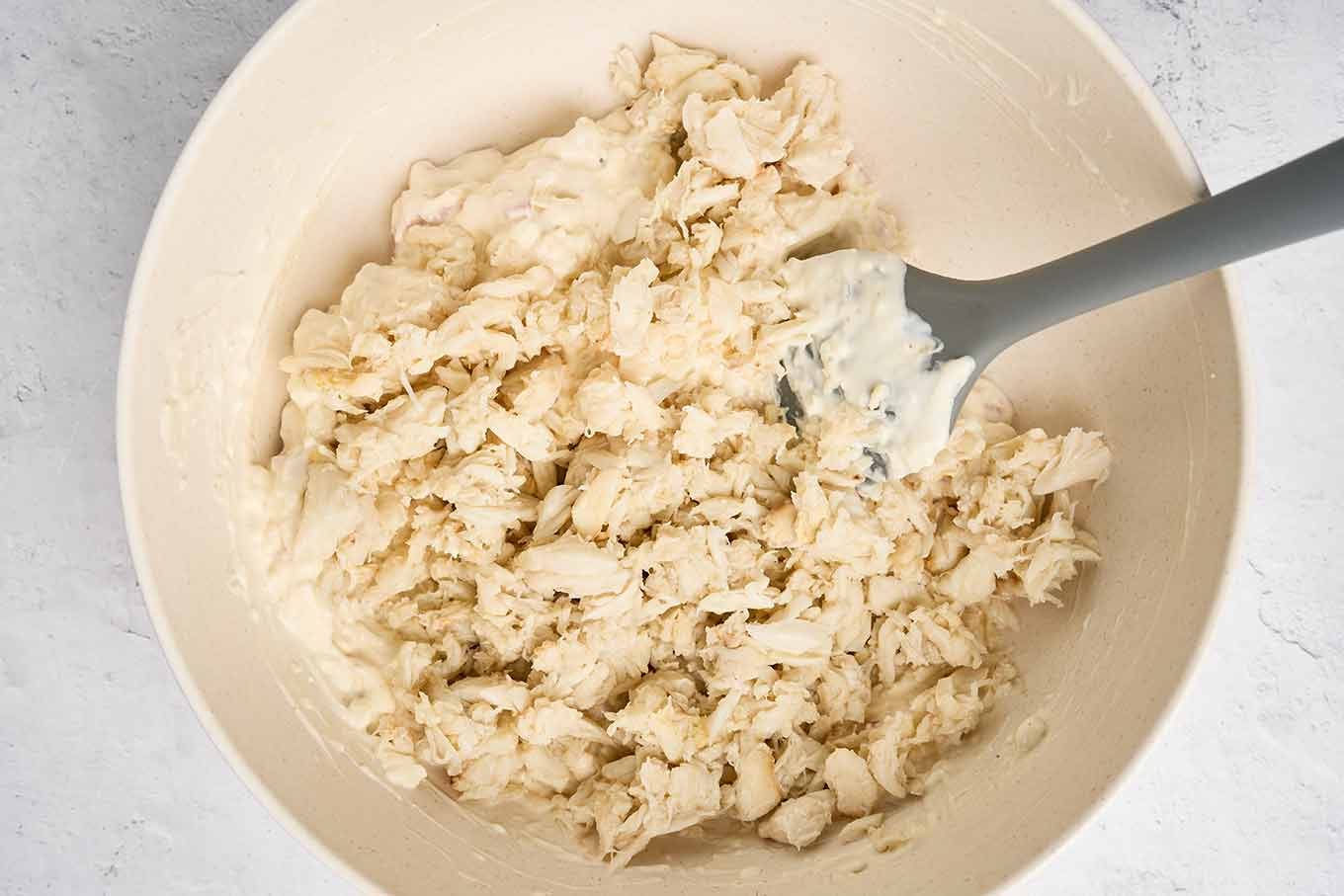 Lump crabmeat being folded into a creamy mixture in a mixing bowl with a spatula.