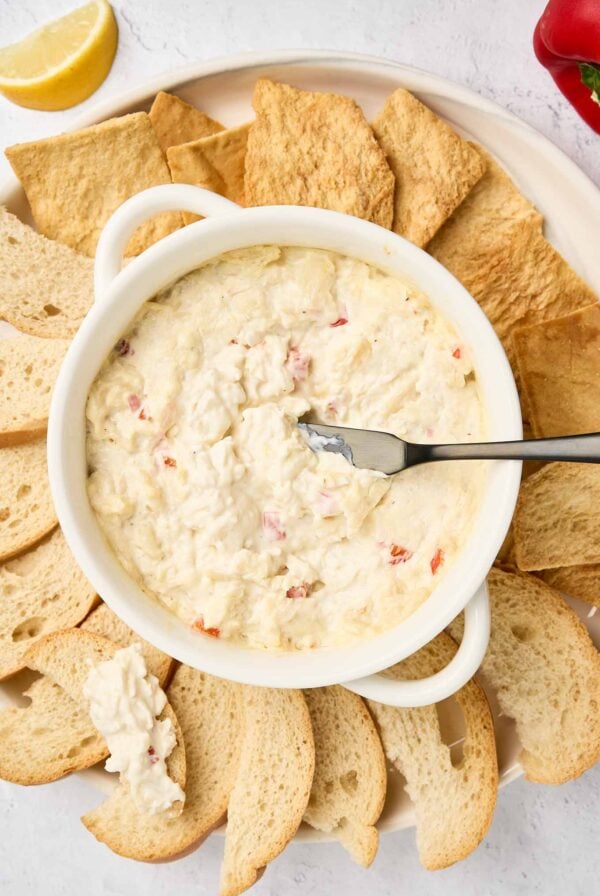 Warm crab dip in a white dish surrounded by toasted bread slices and pita chips, with a spreader resting in the dip.