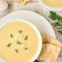 Greek egg and lemon soup garnished with fresh dill, served with pita bread and a soup spoon on a white plate.