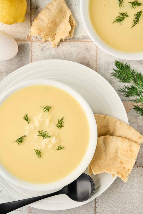 Greek egg and lemon soup garnished with fresh dill, served with pita bread and a soup spoon on a white plate.