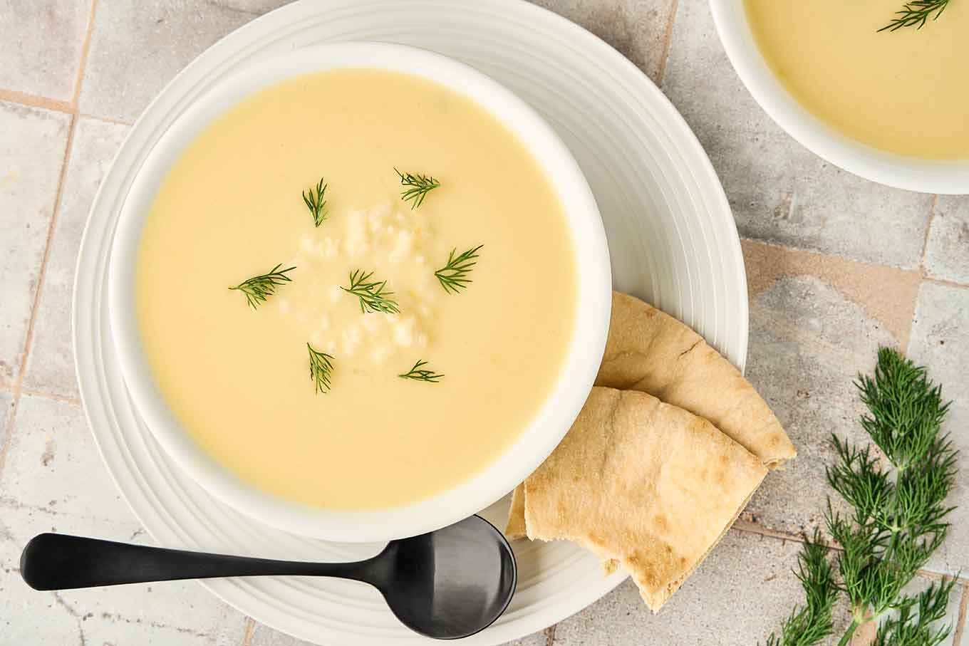 Greek egg and lemon soup garnished with fresh dill, served in a white bowl with pita bread on the side.