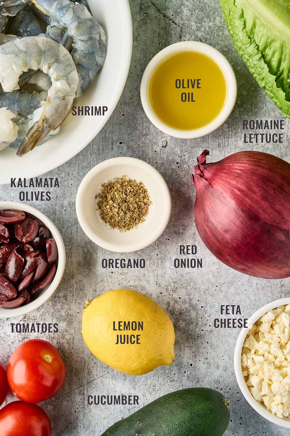 Ingredients for Greek salad with shrimp including raw shrimp, romaine lettuce, tomatoes, cucumber, red onion, feta, olives, lemon juice, olive oil, and oregano.