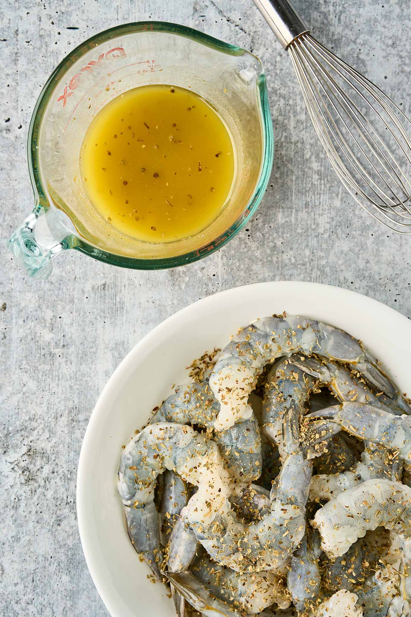 Raw shrimp seasoned with dried oregano in a bowl, with lemon-olive oil dressing and a whisk nearby.