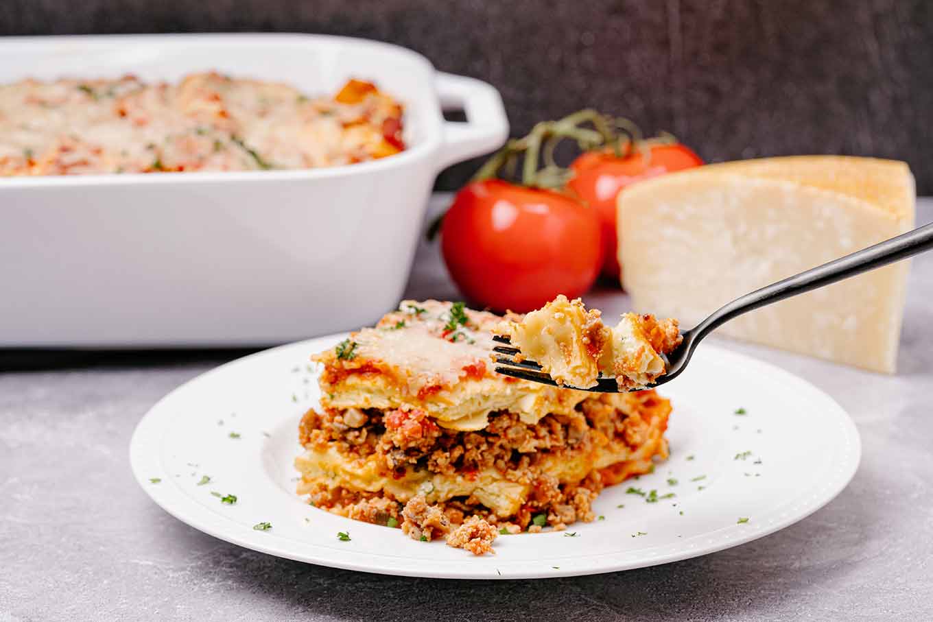 A single serving of lasagna with a forkful being lifted up, the baking dish, fresh tomatoes, and a wedge of cheese in the background.