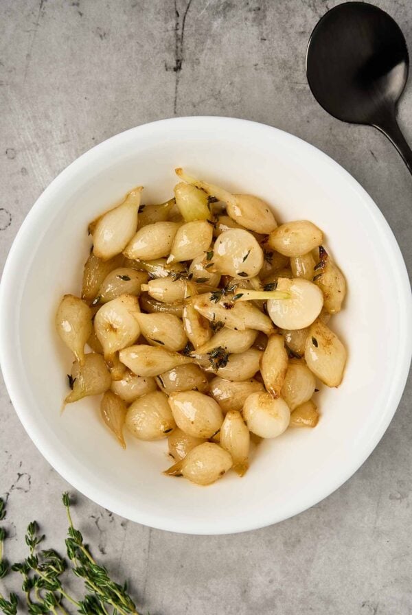 Roasted pearl onions with butter and thyme, lightly caramelized and arranged in a white serving bowl.