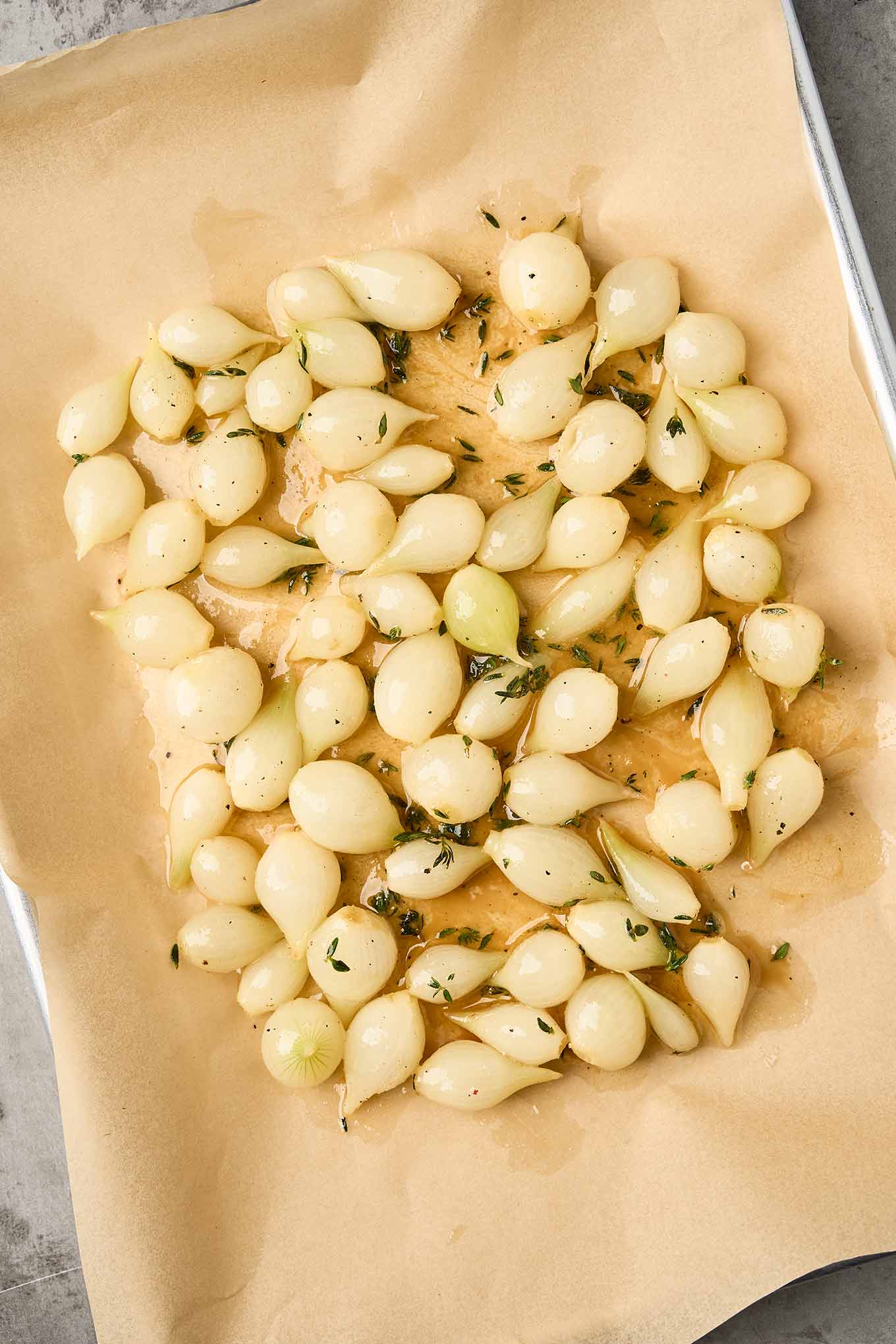 Pearl onions coated with butter, honey, and thyme arranged on parchment paper before roasting.