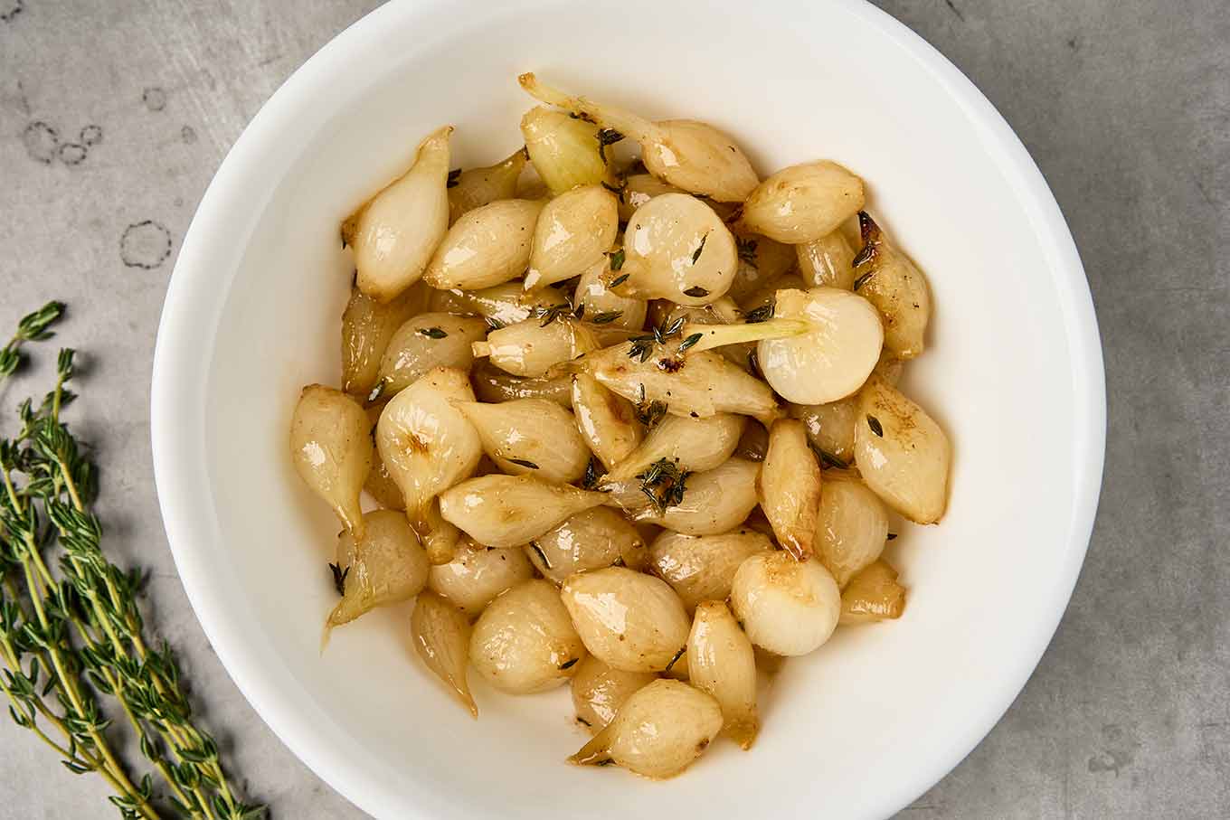 Roasted pearl onions with butter and thyme served in a white bowl - sprigs of fresh time to the side.