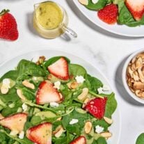 Spinach and strawberry salad with goat cheese, toasted almonds, and creamy balsamic dressing on white plates.