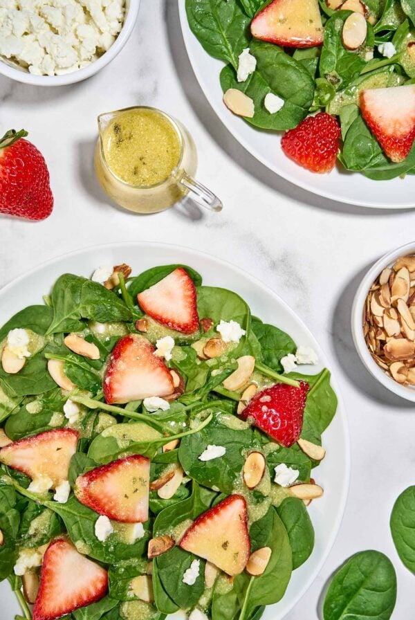 Spinach and strawberry salad with goat cheese, toasted almonds, and creamy balsamic dressing on white plates.