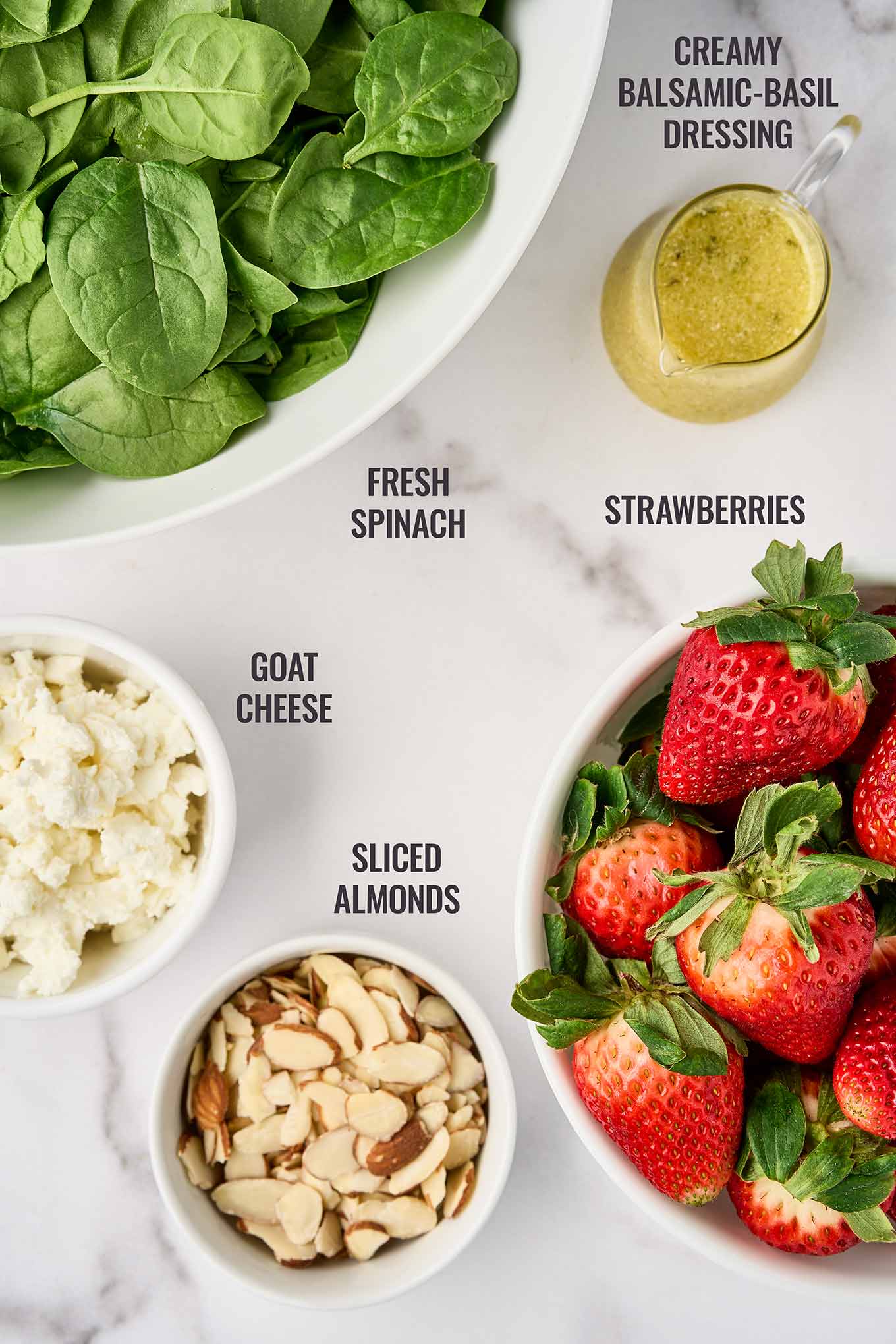 Ingredients for spinach and strawberry salad including baby spinach, strawberries, goat cheese, sliced almonds, and balsamic dressing.