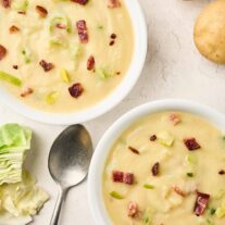 Two bowls of creamy cabbage and potato soup with bacon and leeks, with whole potatoes and cabbage nearby.