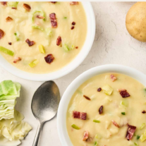Creamy cabbage leek potato soup with bacon and leeks in two bowls, surrounded by whole potatoes, cabbage, and a spoon.