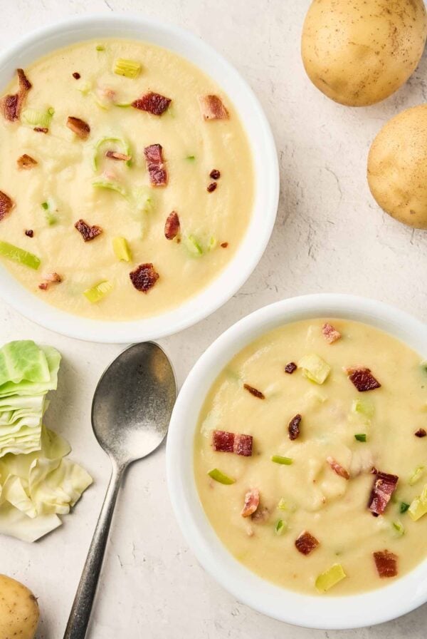 Two bowls of creamy cabbage and potato soup with bacon and leeks, with whole potatoes and cabbage nearby.