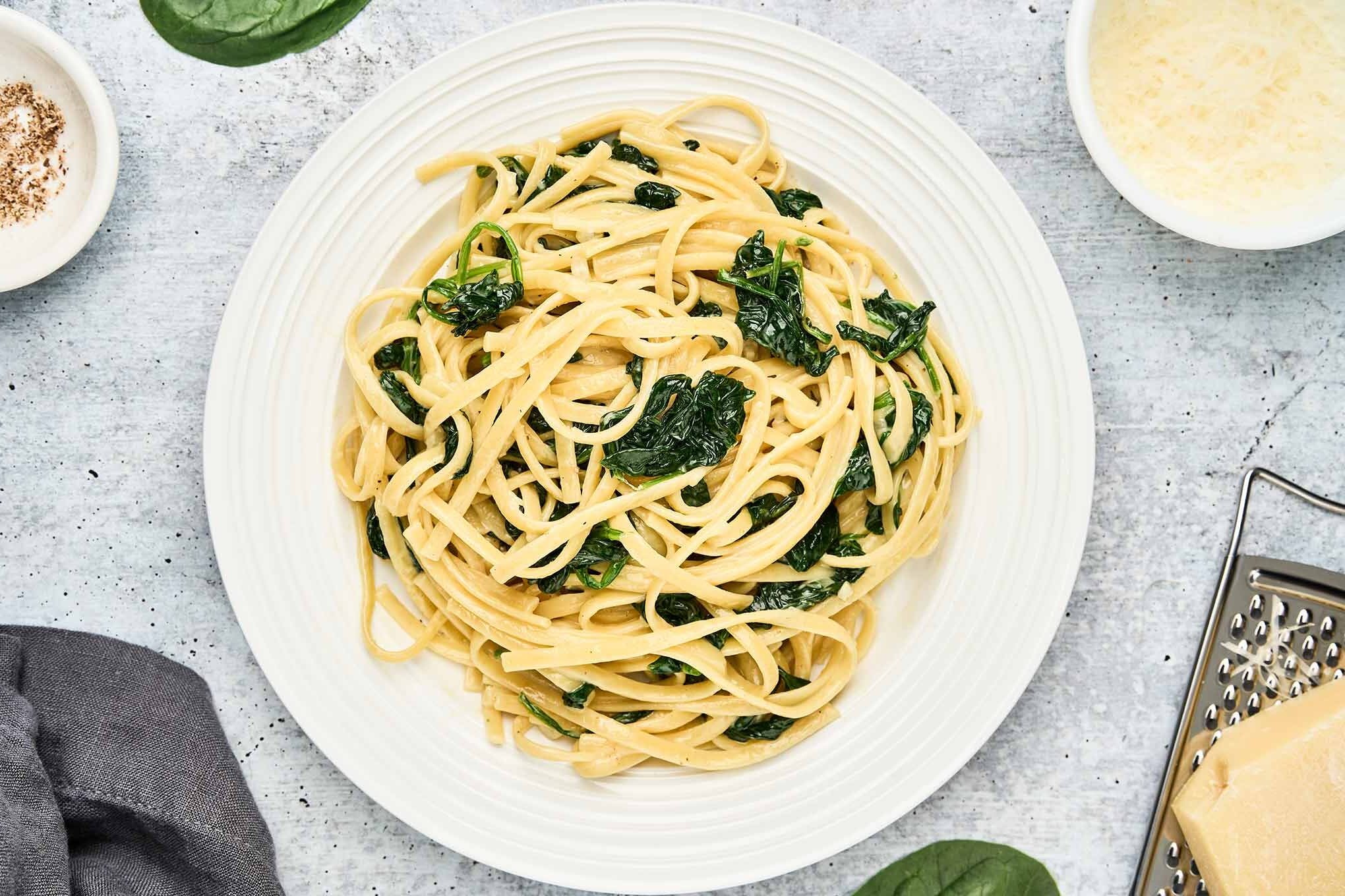 Overhead view of creamy linguine with spinach on a white plate, surrounded by grated Parmesan, a wedge of cheese on a grater, fresh spinach leaves, nutmeg, and a gray napkin on a textured surface.