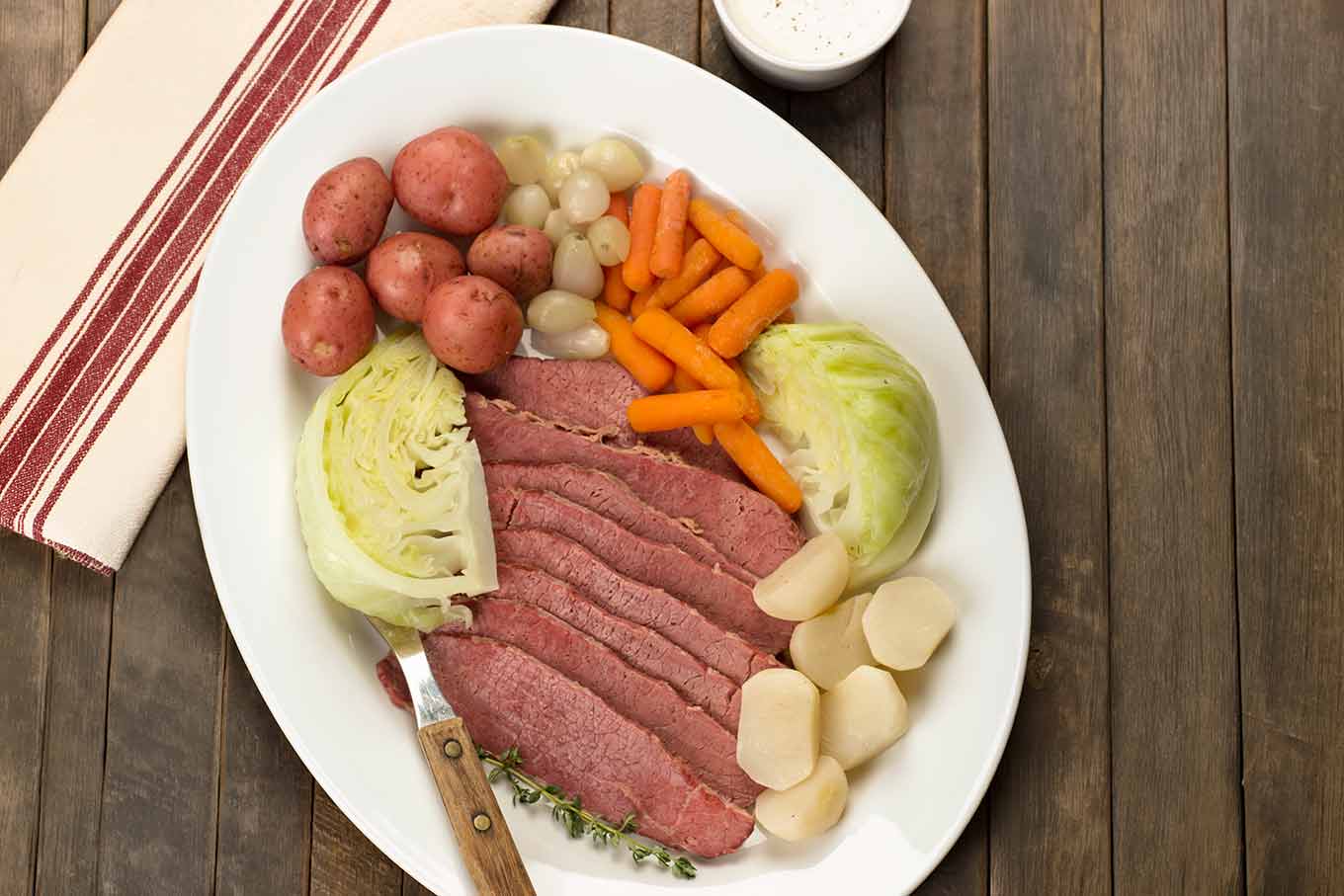 Sliced corned beef brisket arranged with cabbage wedges, baby potatoes, carrots, turnips, and pearl onions on a white platter.
