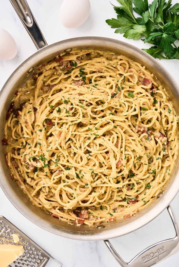 One-pot spaghetti carbonara with bacon, Parmesan, and parsley in a stainless skillet, eggs and cheese visible alongside.