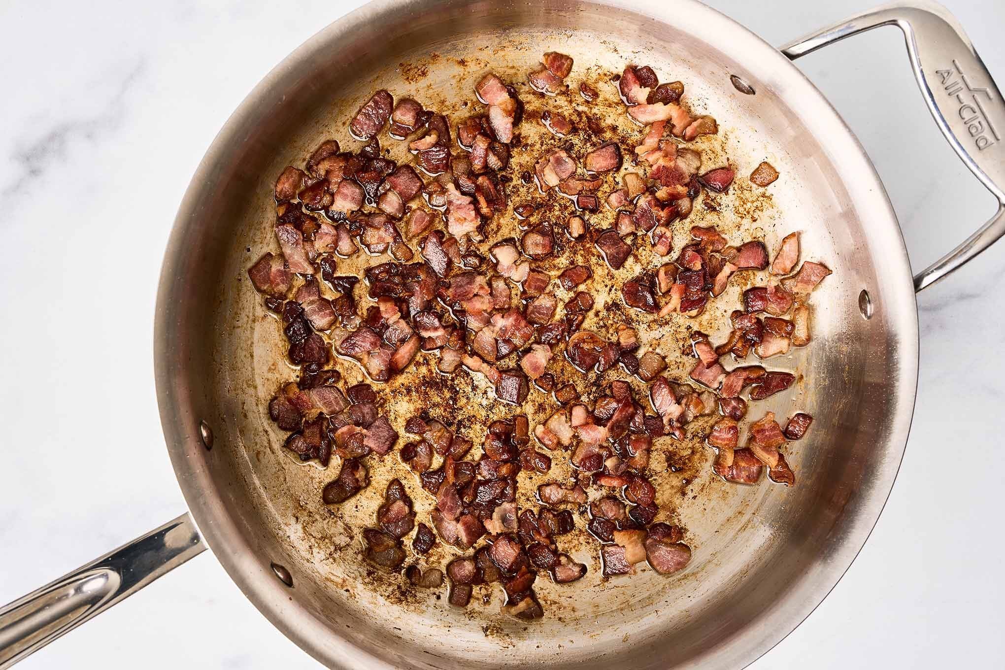 Bacon pieces cooking in a stainless skillet until crisp with rendered fat and browned bits in the pan.