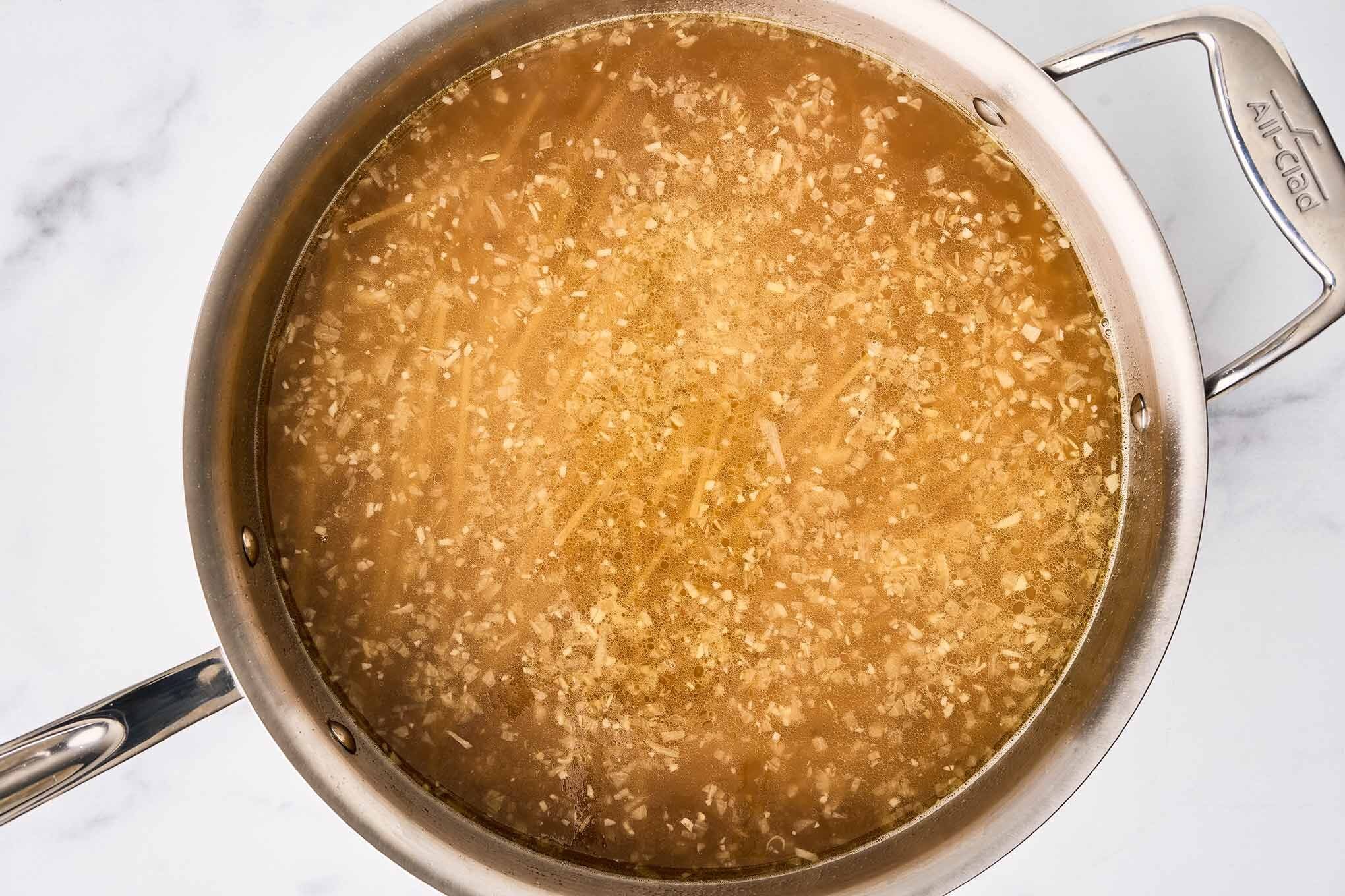Thin spaghetti cooking in chicken broth with saut&eacute;ed garlic and shallot in a stainless skillet.