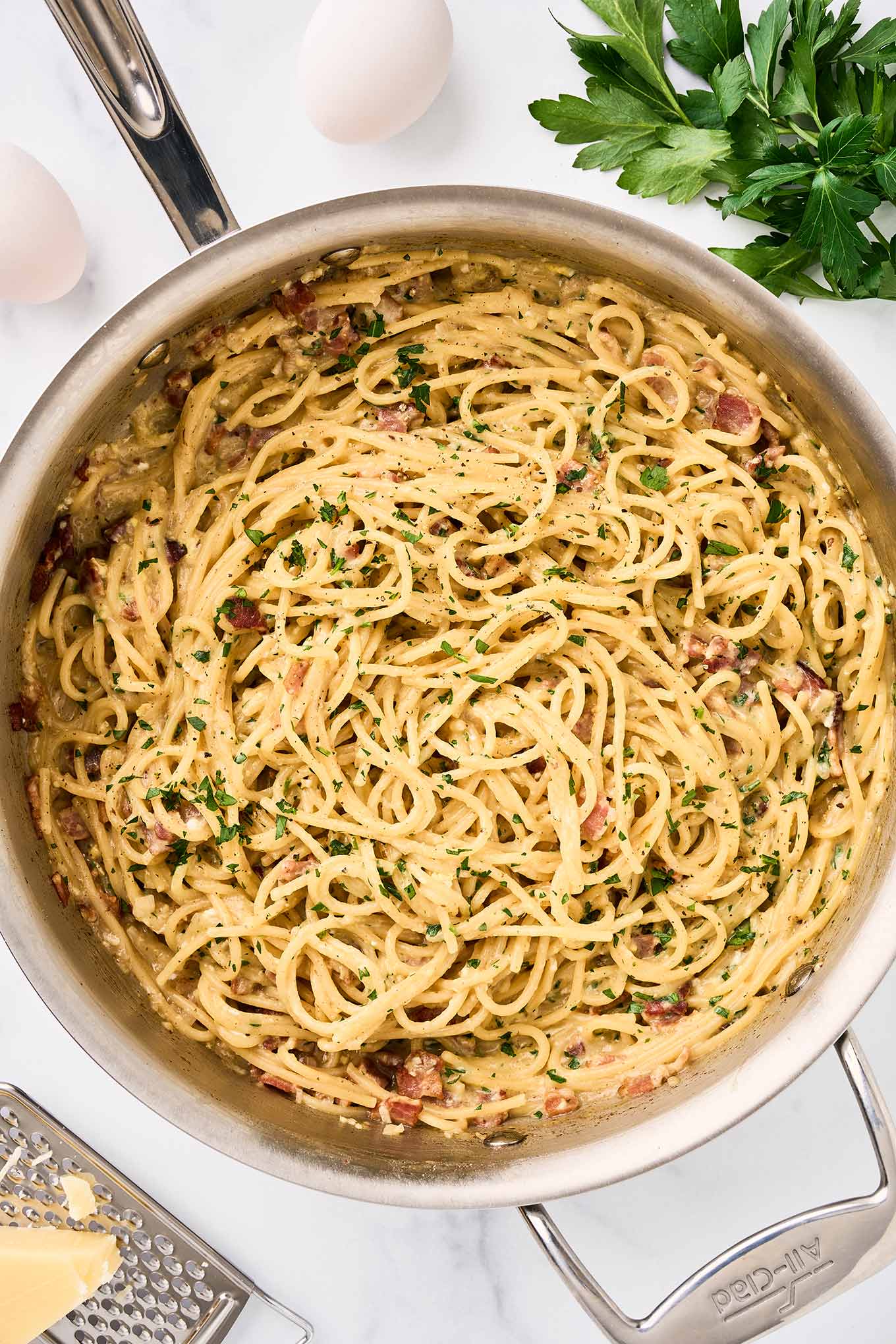 One-pot spaghetti carbonara with bacon, Parmesan, and parsley in a stainless skillet, eggs and cheese visible alongside.