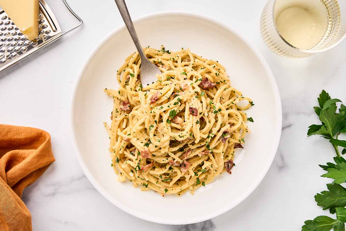 A serving of one-pot spaghetti carbonara on a white plate with fork, cheese grater and cheese and white wine on the side.