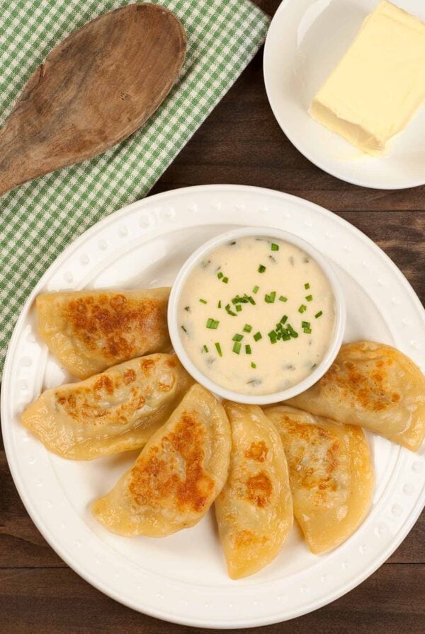 Homemade pierogi with potato and cheese filling, pan-fried until golden, served with sour cream chive sauce on a white plate.