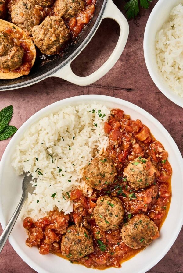 Moroccan meatballs in tomato sauce served over white rice on a plate, with extra meatballs in a skillet and fresh herbs garnish.