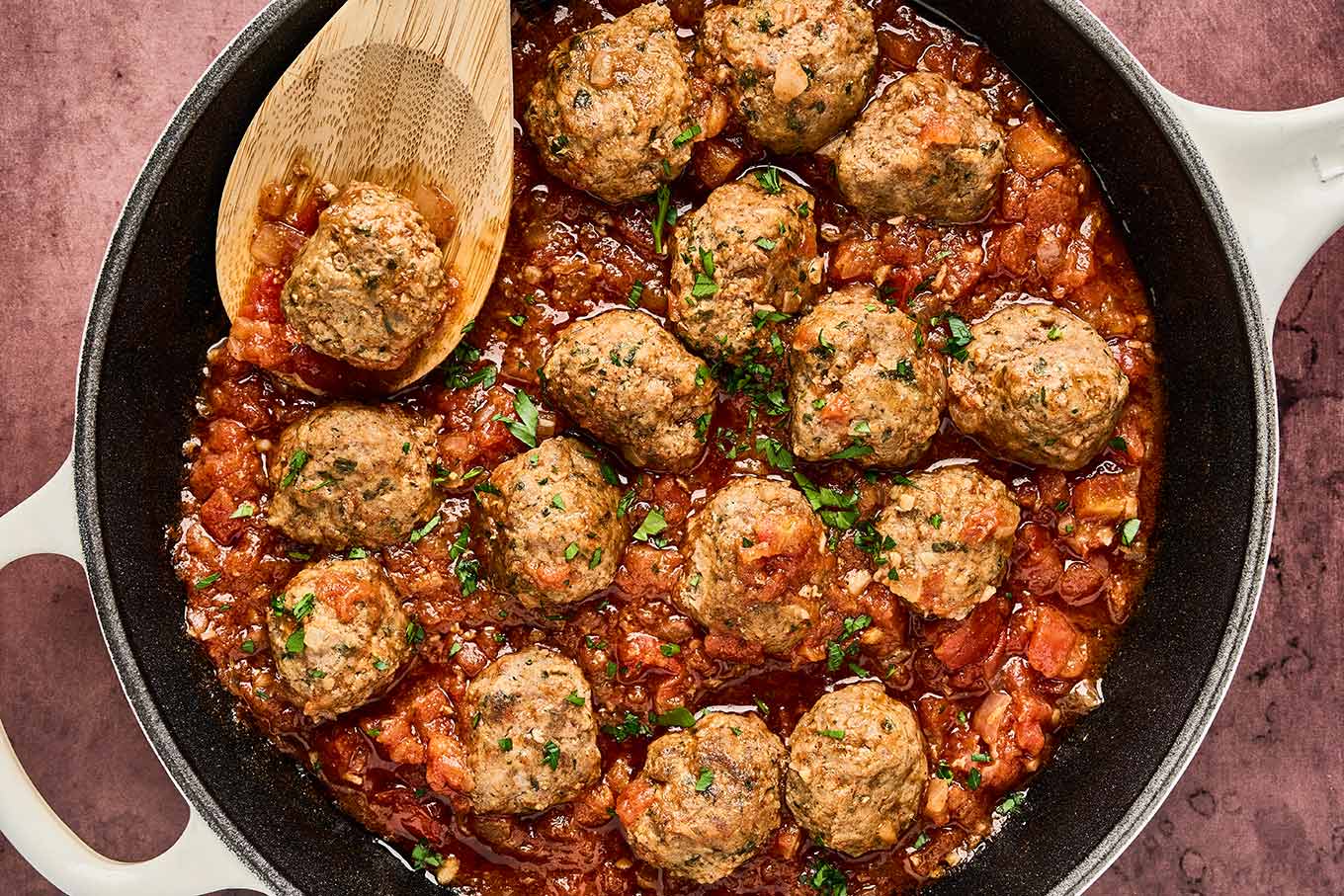 Moroccan meatballs simmering in tomato sauce in a skillet, with chopped herbs and wooden spoon.