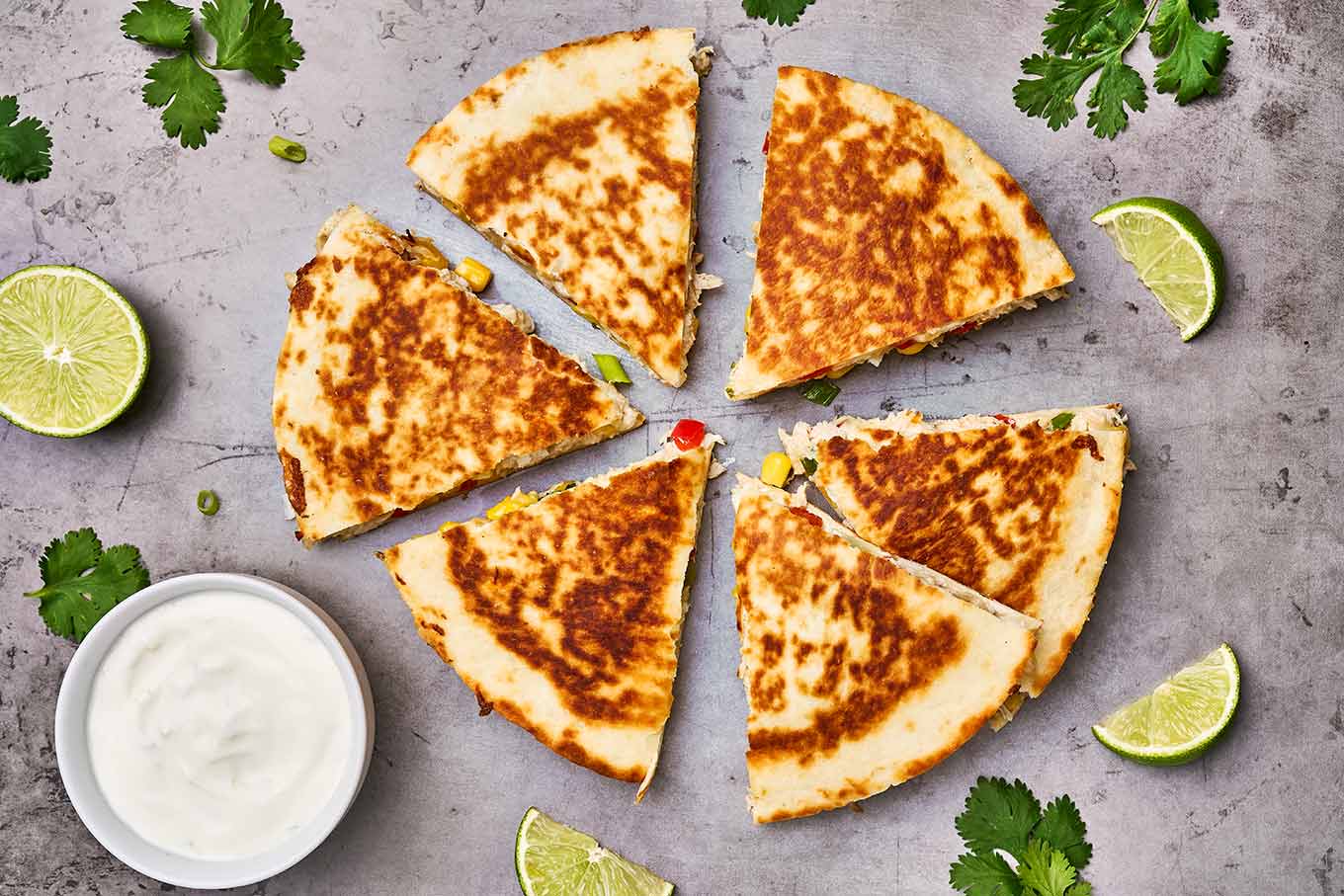 Toasted chicken quesadilla cut into wedges, surrounded by lime wedges and a bowl of lime crema on the side.