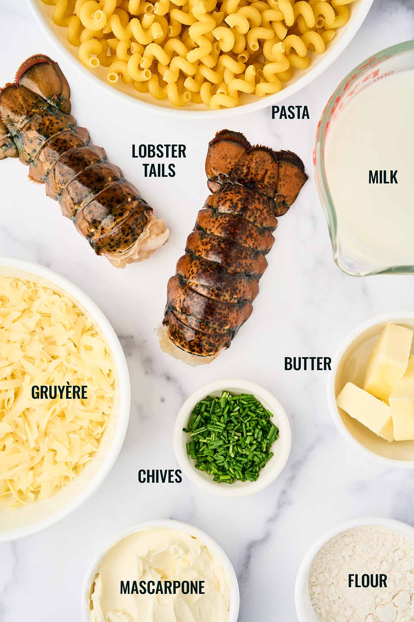 Ingredients for stovetop lobster mac and cheese including lobster tails, cavatappi pasta, Gruy&egrave;re, mascarpone, milk, butter, flour, and chives.