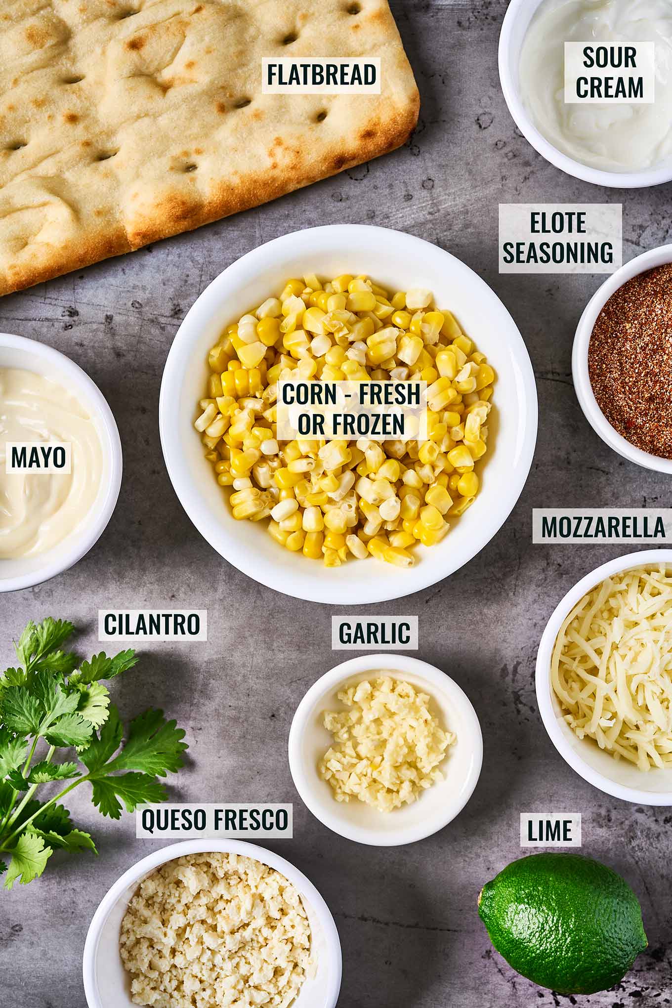 Ingredients for Mexican street corn flatbread; flatbread, sour cream, corn, elote seasoning, mayo, mozzarella, garlic, queso fresco, lime, and cilantro.