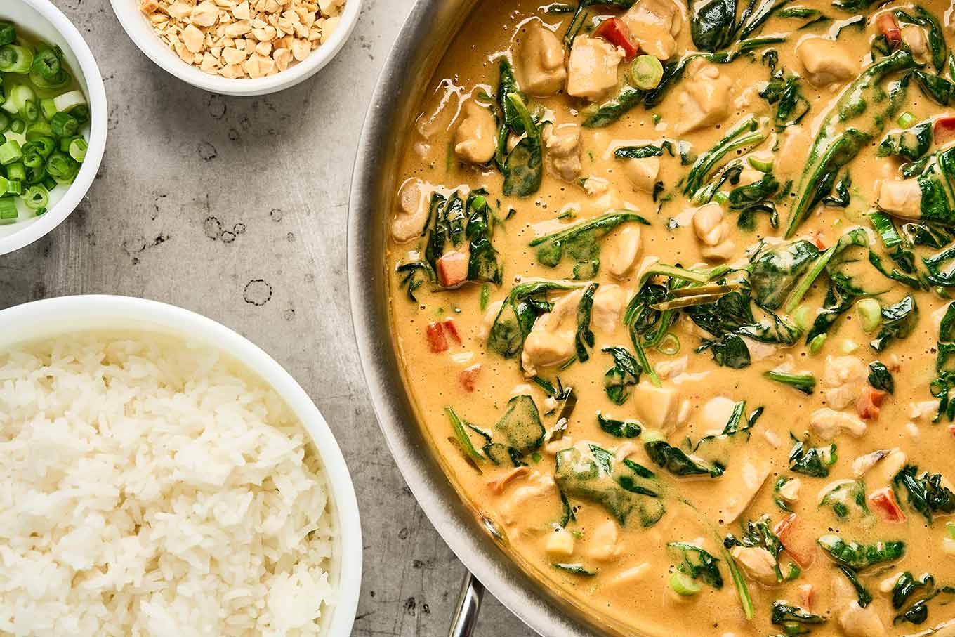 Thai peanut curry with chicken and spinach in a sauté pan beside jasmine rice, scallions, and chopped peanuts.