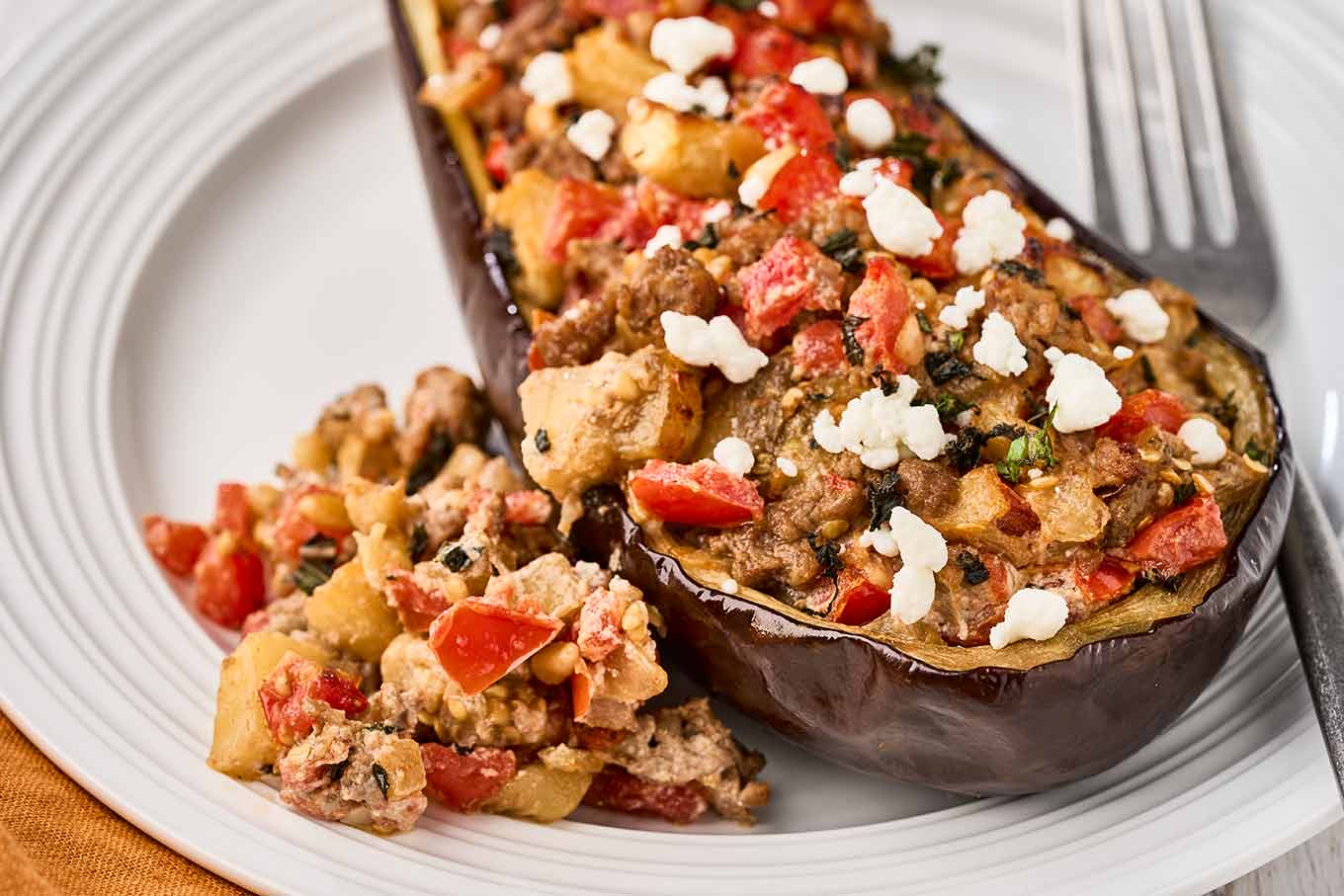 Mediterranean stuffed eggplant with lamb, tomatoes, pine nuts, and goat cheese on a white plate.